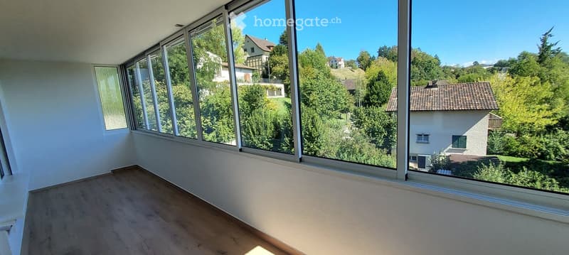 Empty room with large windows offering a scenic view of trees and houses. The floor is wooden, and the walls are white.