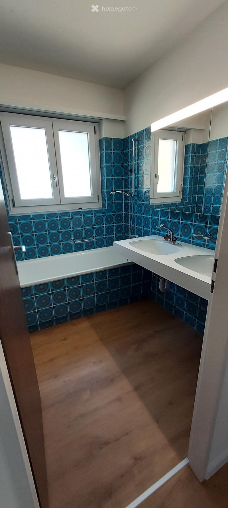 Bathroom with blue patterned tiles, a white bathtub, a double sink, and a mirror above the sink. The floor is wooden.