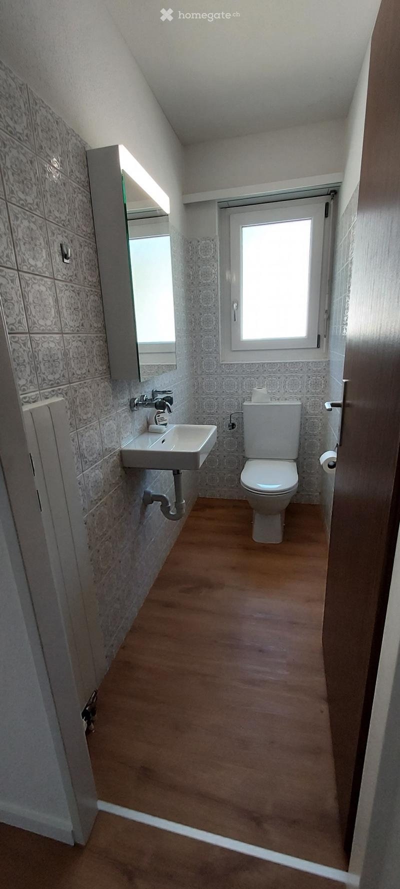 Modern bathroom with wooden flooring, white sink, toilet, and a mirror above the sink. The walls are tiled with a decorative pattern, and there is a window providing natural light.