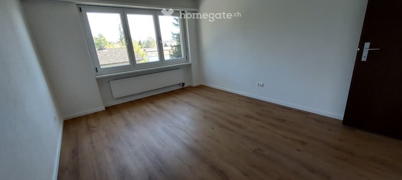 Empty room with wooden flooring, white walls, large window, radiator, and a closed door.