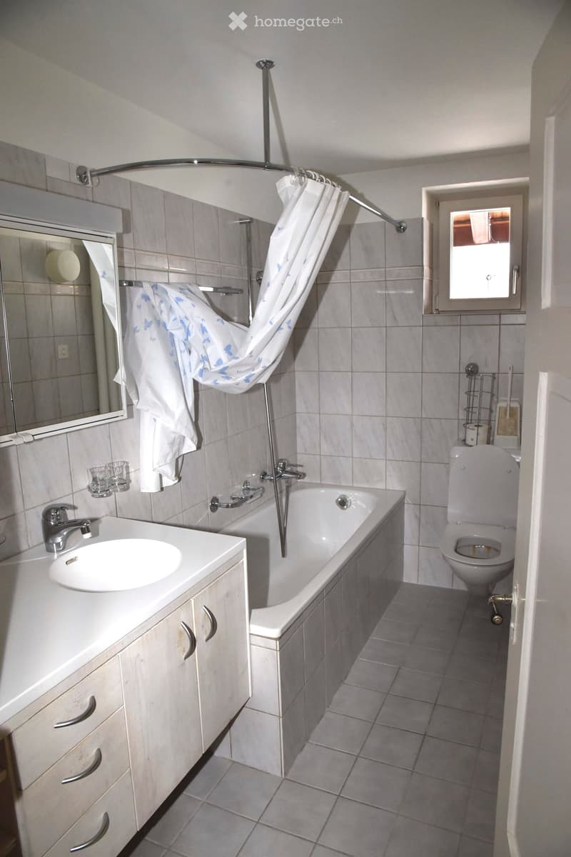 Bathroom with a white sink, bathtub, toilet, mirror, and a shower curtain. The room features tiled walls and floor, and a small window letting in natural light.