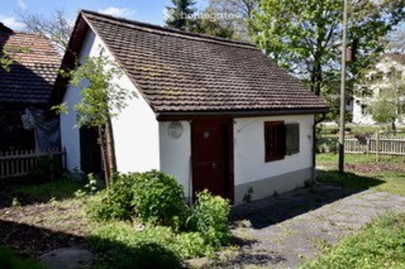 A small, single-story house with a brown tiled roof, white exterior walls, and a wooden front door. The house is surrounded by a small garden area with shrubs and a tree, and is situated on a paved driveway.