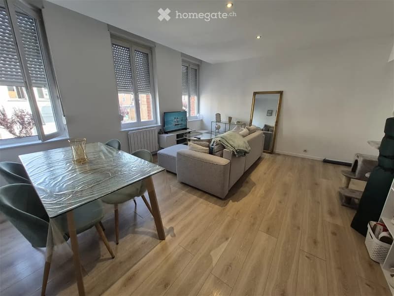 Modern living room with light wooden flooring, large windows, a grey sofa, a glass dining table with chairs, a TV stand, and a decorative mirror.