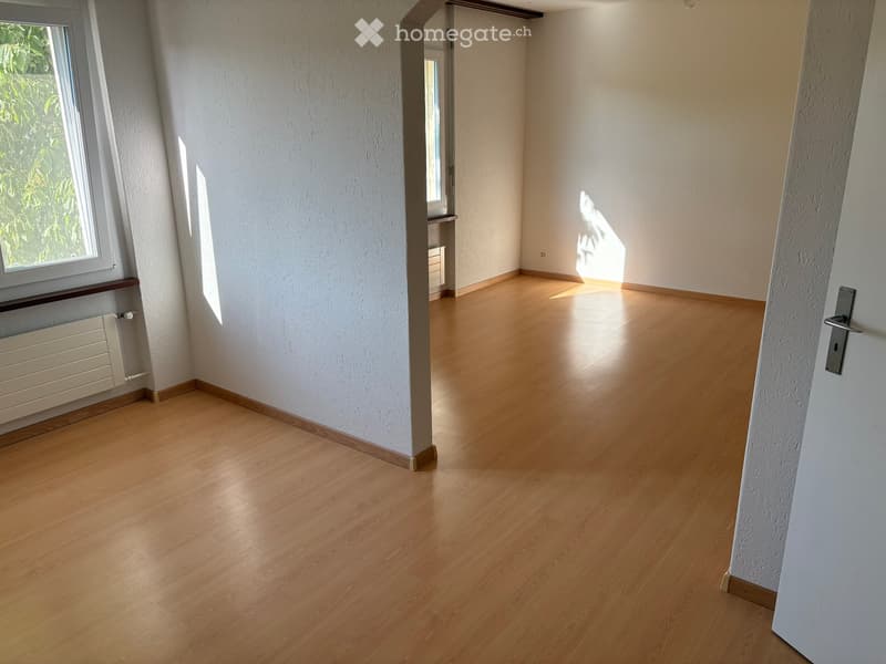 Empty room with light wooden flooring, white walls, and large windows allowing natural light to enter.