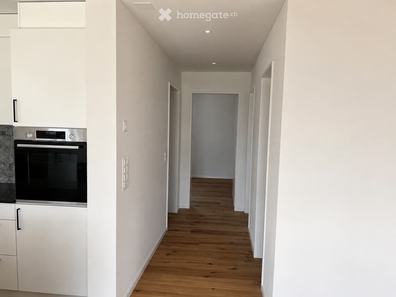 A narrow, well-lit hallway with wooden flooring and white walls, leading to an open doorway. The hallway features recessed ceiling lights and a built-in oven on the left side.