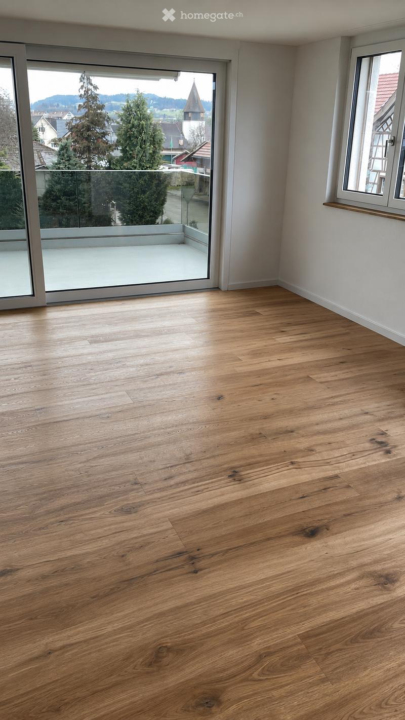 Empty room with light wooden flooring, large windows, and a sliding glass door leading to a balcony.