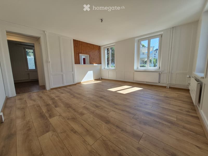 Empty room with wooden flooring, white walls, and large windows allowing natural light. There is a doorway leading to another room with wooden paneling.