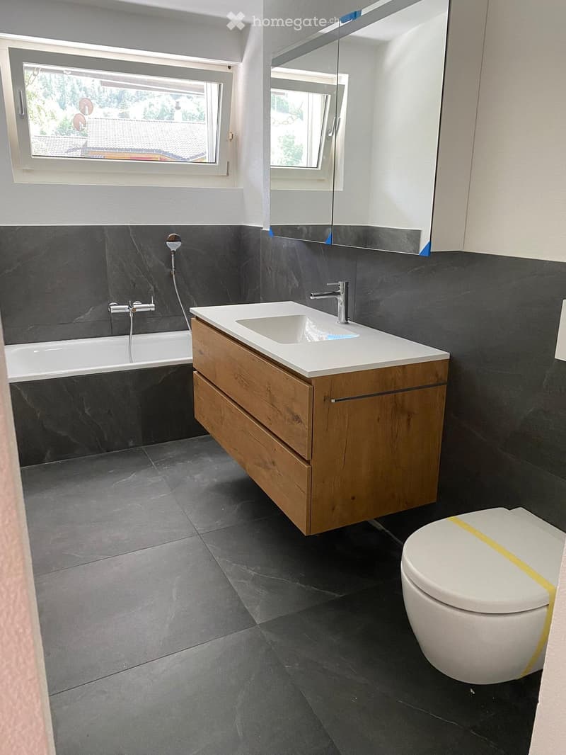 Modern bathroom with dark grey tiles, a white bathtub, a wooden sink cabinet, a mirror, and a toilet.