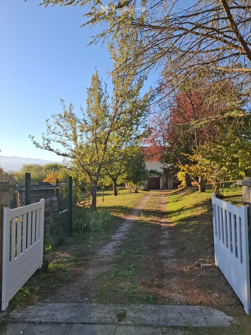 A gated entrance leads to a pathway surrounded by trees and greenery, with a house visible in the background.