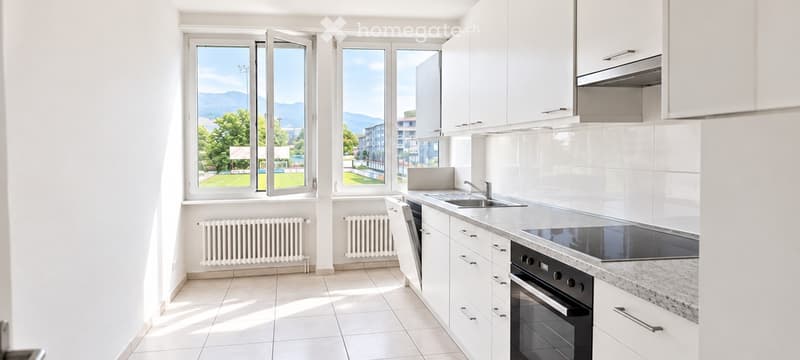 Modern, well-lit kitchen with white cabinets, a countertop, sink, oven, and dishwasher. Large windows offer a scenic view of the outdoors.