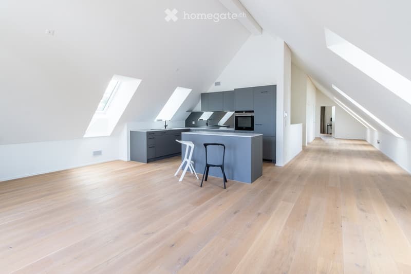 Modern kitchen with grey cabinets, a central island, and light wooden flooring. Two stools and a chair are placed near the island, and the space features skylight windows allowing natural light to fill the room.