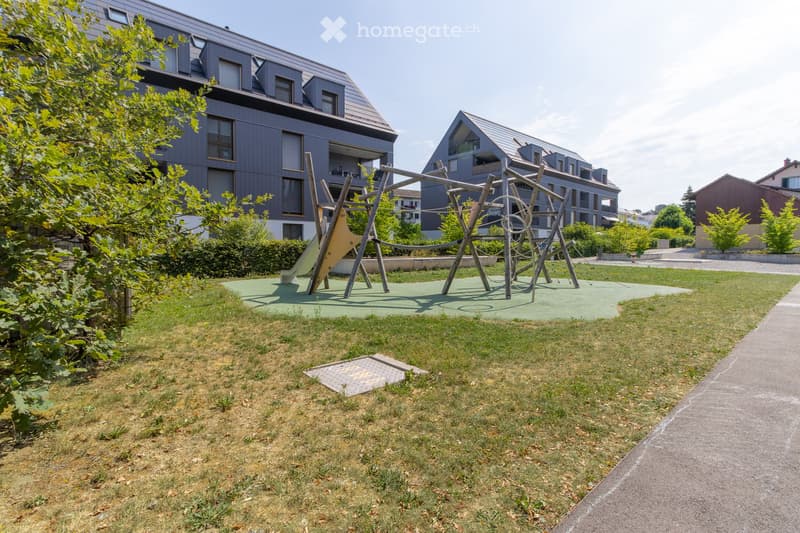 Outdoor playground area with a slide, climbing structure, and grassy lawn, surrounded by modern apartment buildings.