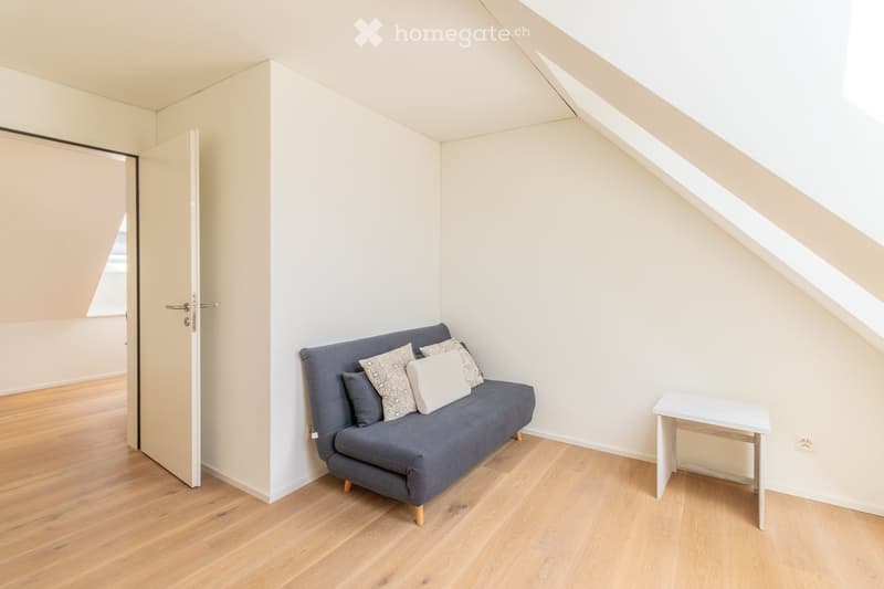 A small, modern attic room with wooden flooring, white walls, a grey sofa with decorative pillows, and a small white side table.