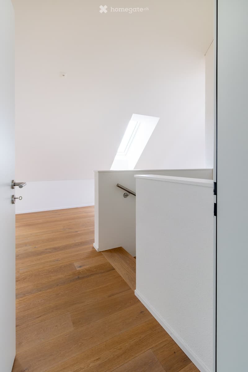 Empty room with light wooden flooring, white walls, and a skylight window providing natural light. The room features a door with a silver handle and a small white partition wall with a built-in shelf and a handrail.