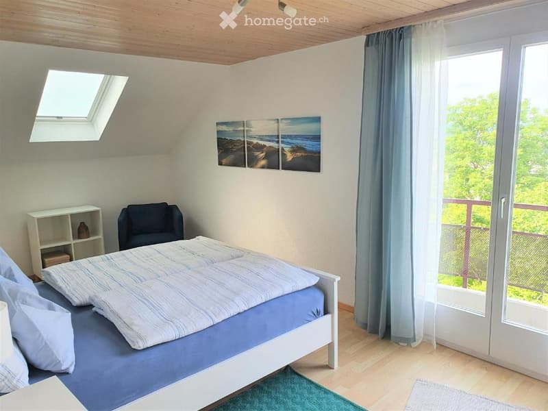 Cozy bedroom with a bed, a chair, a small shelf, and a large window leading to a balcony. The room features a slanted wooden ceiling and a skylight.