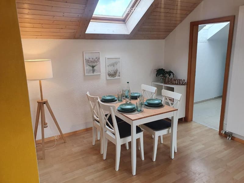 Bright dining area with wooden floor, skylight, white walls, dining table with six chairs, floor lamp, picture frames, and a small welcome sign with a plant on a shelf.