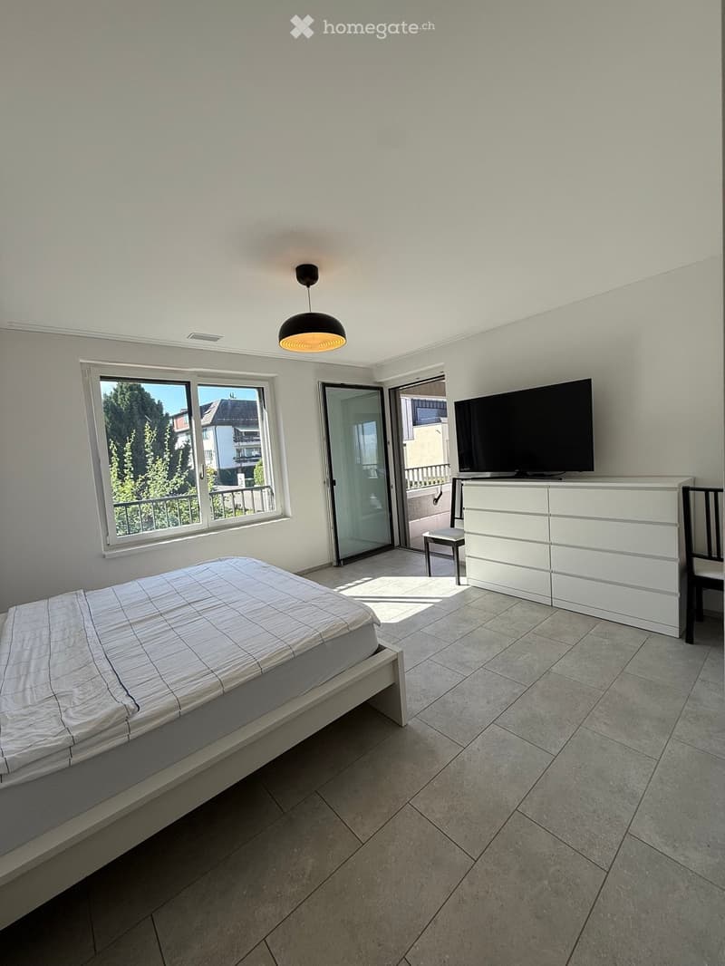 Modern bedroom with a white bed, dresser, chair, and TV. Large window with a view and a sliding glass door leading to a balcony.