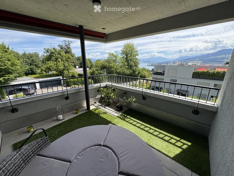 Balcony with a view of mountains and cityscape, featuring a wicker chair, small table, potted plants, and artificial grass.