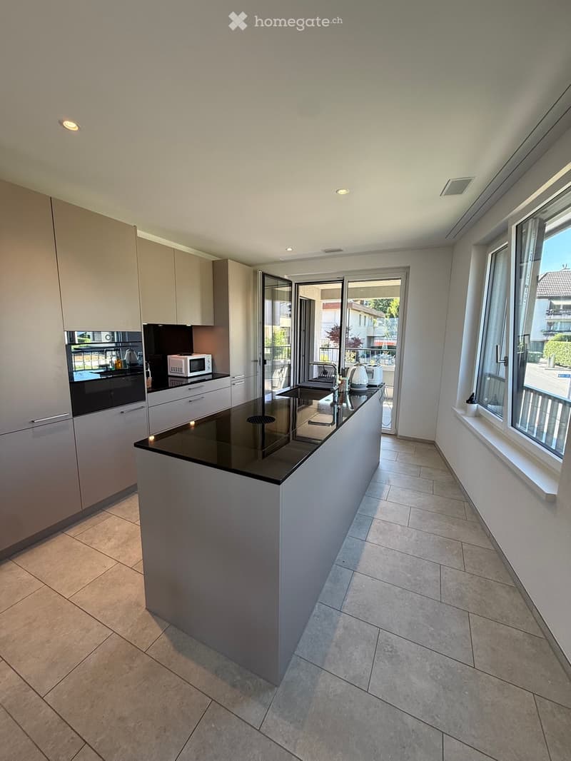 Modern kitchen with a central island, built-in microwave, oven, sink, and ample cabinetry. Large windows and sliding doors lead to a balcony.