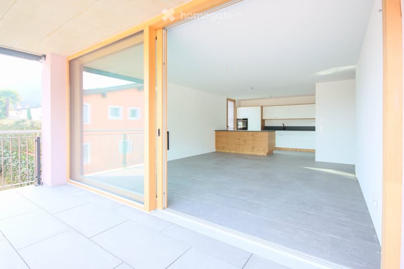 Open-plan kitchen and living area with modern wooden kitchen island, white cabinets, and grey tiled flooring. Large sliding glass doors lead to a tiled balcony with metal railing.