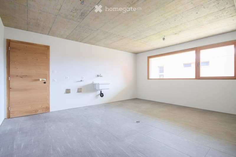 Empty room with white walls, grey tiled floor, wooden door, and a small sink with a faucet. The ceiling features a mix of exposed stone and wooden panels, and there are two large windows with wooden frames allowing natural light to enter.
