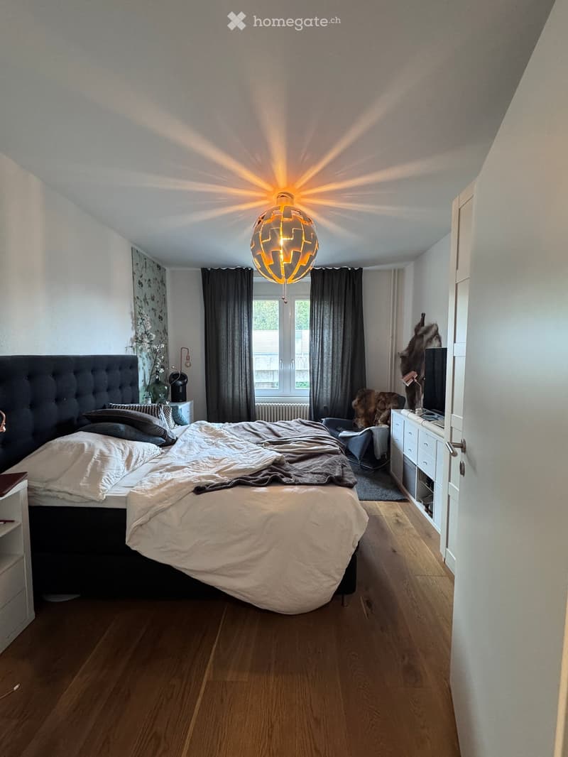 Cozy bedroom with wooden flooring, large bed with white bedding, nightstand with lamp, window with dark curtains, and a pendant light fixture.