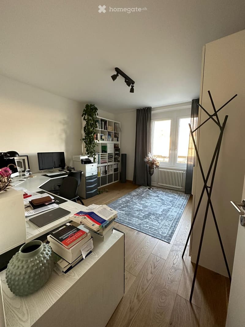 A well-lit study or home office with a wooden floor, large window with curtains, desk with laptop, books, and a vase, a bookshelf with books and a plant, and a coat rack near the door.