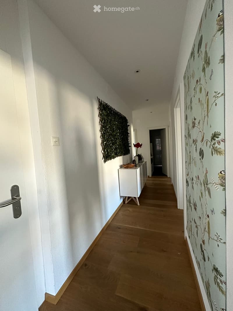 A narrow hallway with wooden flooring, white walls, and a decorative mural featuring birds and leaves. A small white console table with a vase and a plant is visible on the left side.