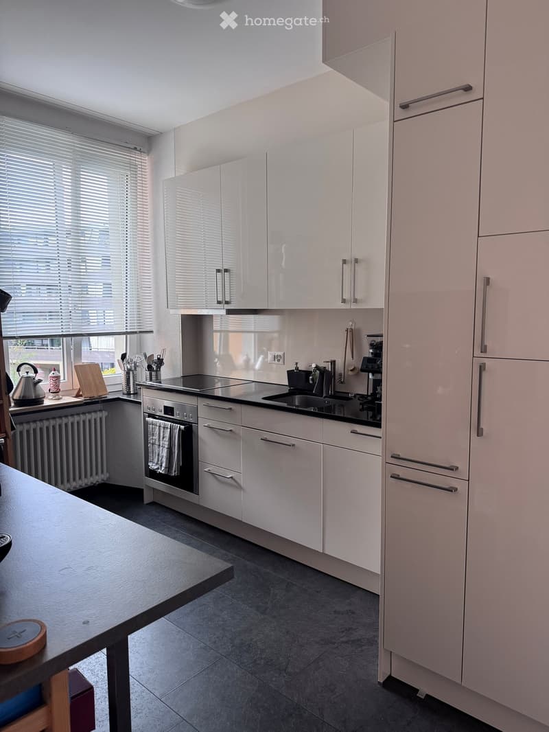 Modern kitchen with white cabinets, stainless steel appliances, and a dark countertop. Features an oven, sink, coffee maker, and various kitchen utensils.