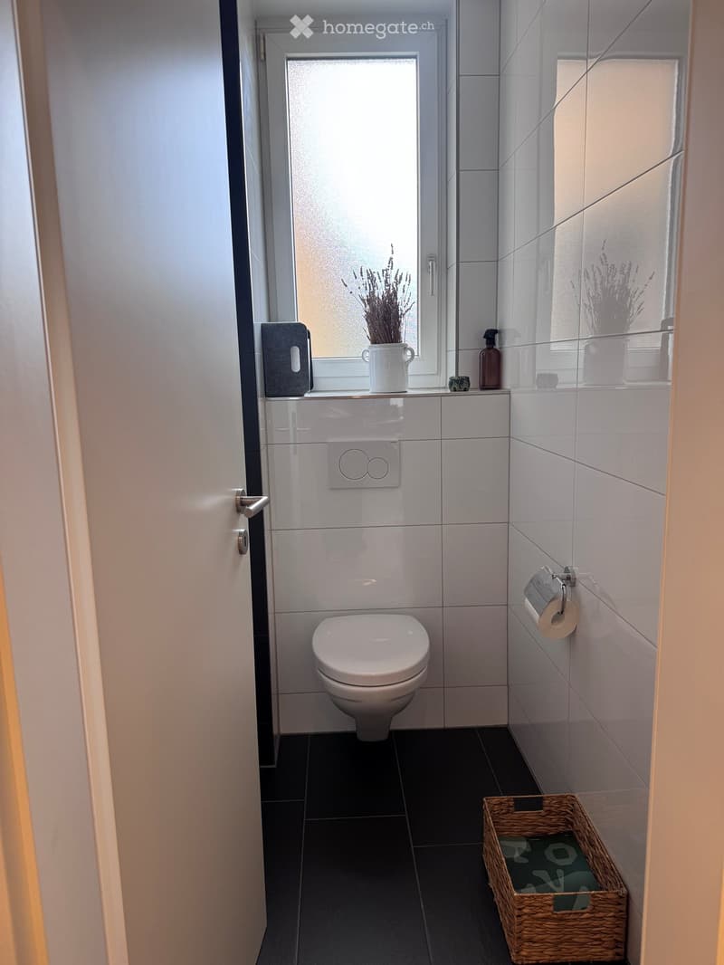 Modern bathroom with white tiles, black floor, toilet, toilet paper holder, wicker basket, vase with dried flowers, and bottle on windowsill.