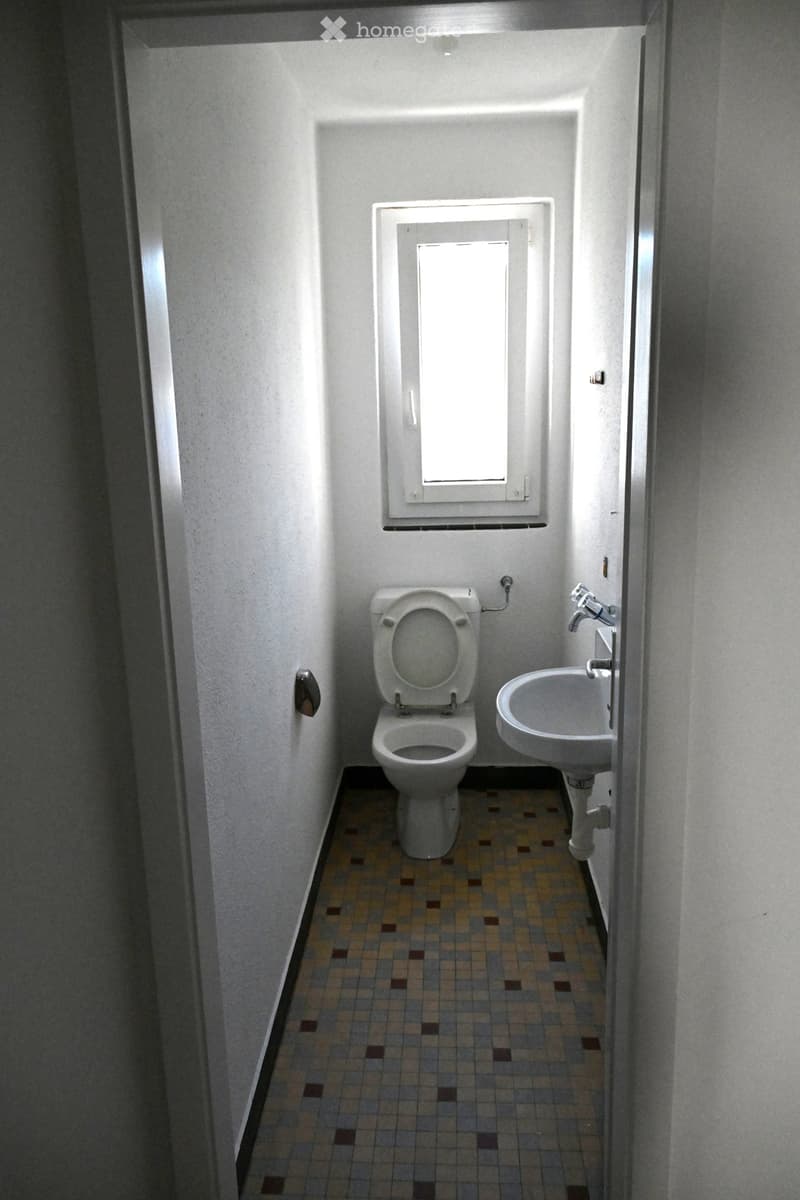 Small bathroom with a toilet, sink, and a window above the toilet. The floor is tiled with a pattern of brown and beige tiles.