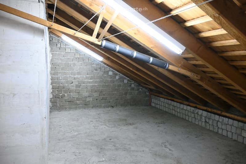 Empty attic space with exposed wooden beams, concrete floor, and brick walls. No furniture or equipment present.