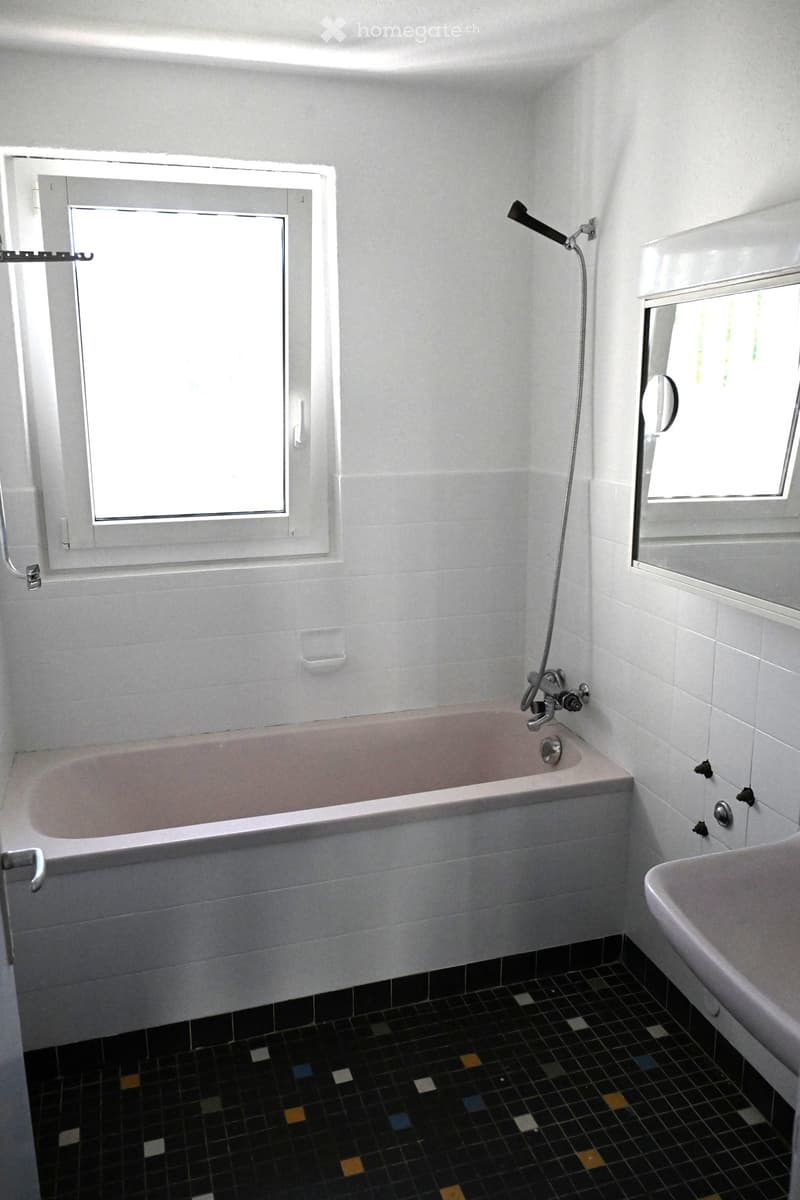 Bathroom with a bathtub, showerhead, sink, and mirror. The walls are tiled in white with a black and colorful mosaic tile floor.