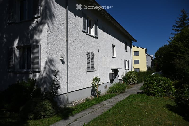 Two-story house with white exterior, multiple windows with shutters, small garden area with pathway, and neighboring yellow building visible.