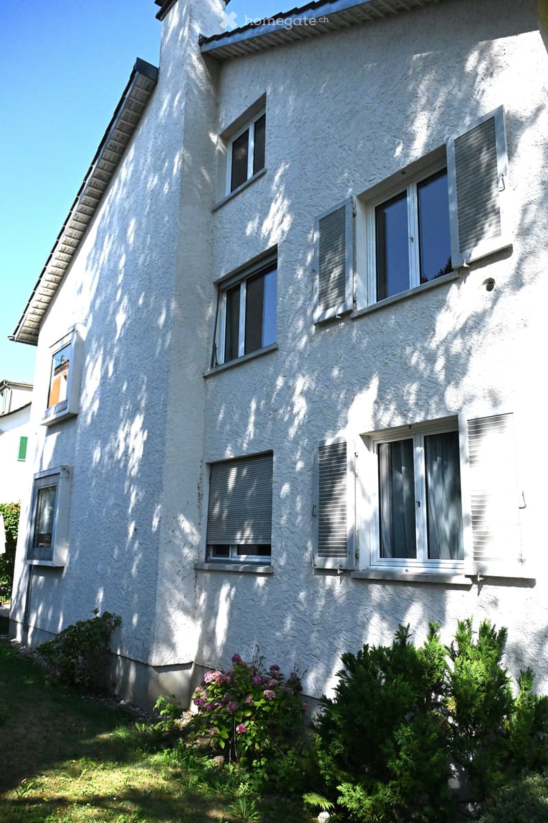 Two-story house with white textured exterior, multiple windows with shutters, and small garden area with shrubs and flowering plants.