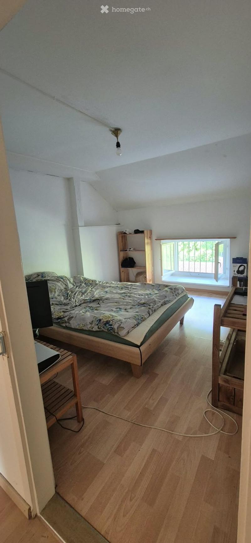 Bright bedroom with wooden flooring, slanted ceiling, and large window with balcony access. Features a wooden bed with patterned bedding, a nightstand, a small shelf, and a laptop on a wooden stand.