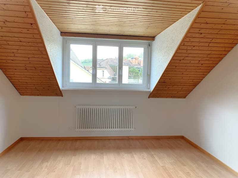 An empty room with wooden floors and wooden ceiling, a large window, and a white radiator