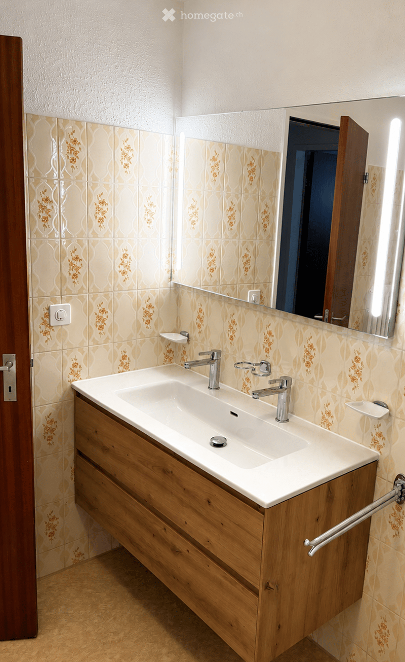 Modern bathroom with a white sink, wooden cabinet, and large mirror with built-in lighting. The walls are tiled with a floral pattern, and the floor is light-colored.