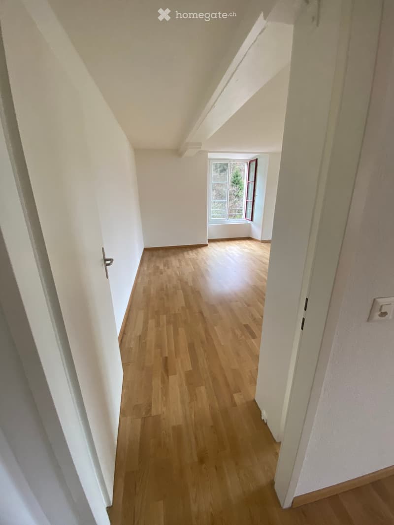 Empty hallway with wooden flooring, white walls, and a window at the end.