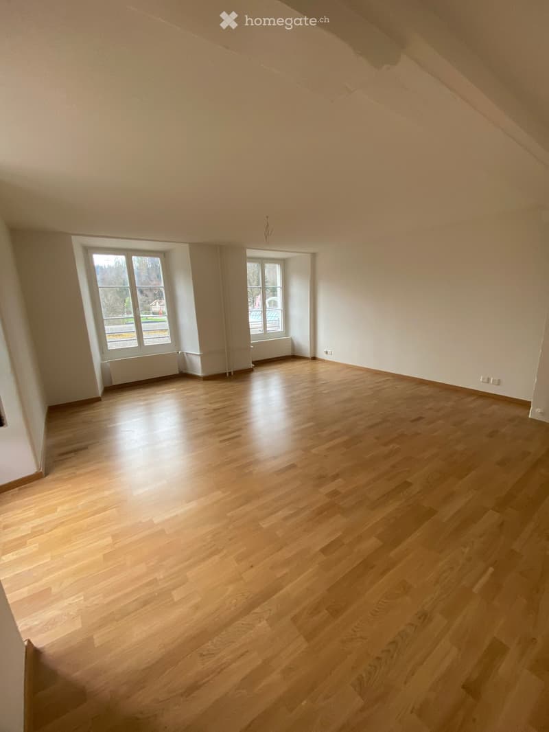 Empty room with wooden flooring and large windows, featuring white walls and radiators.