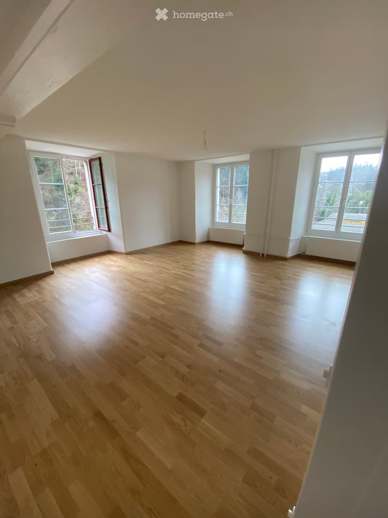 Empty room with wooden flooring, white walls, and large windows with views of greenery outside.