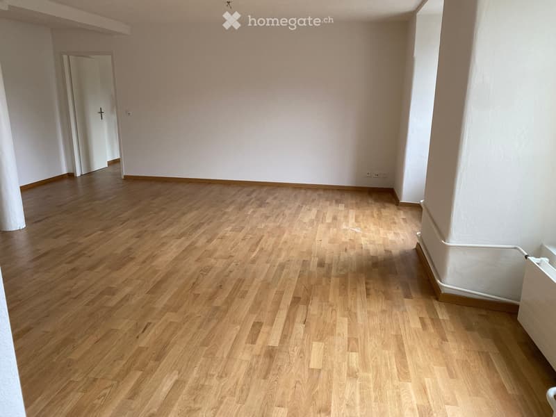 Empty room with light wooden flooring and white walls, featuring a doorway on the left side.