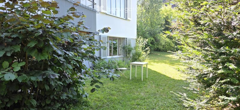A well-maintained garden with a small white table and a chair, surrounded by lush greenery and a white building in the background.
