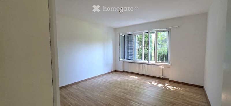 Empty room with wooden flooring, white walls, and large windows with open shutters. A radiator is visible under the windows.