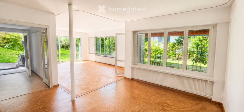 Bright, open room with large windows and wooden flooring, leading to a garden area.