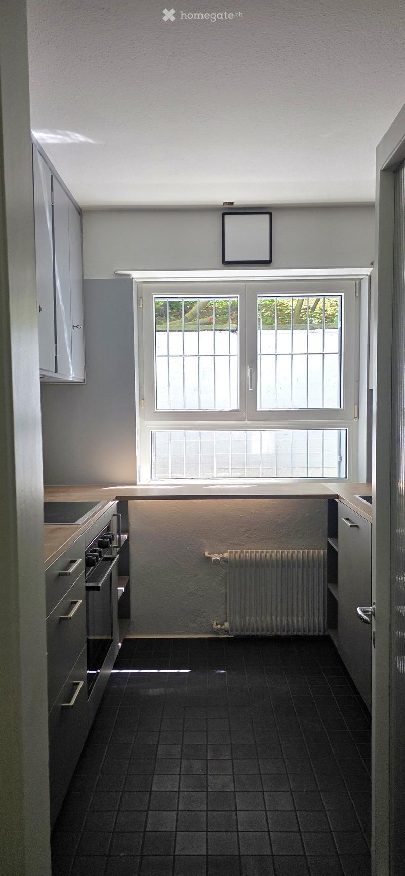 Modern kitchen with black tiled floor, grey cabinets, stove, radiator, and large window with white frame.
