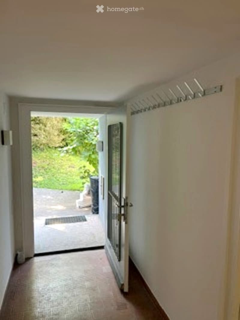 A narrow hallway with wooden flooring, white walls, and an open door leading to a garden area. A coat rack is visible on the right side.