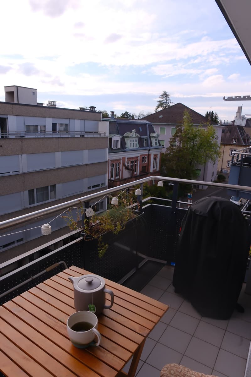 Balcony with wooden table, teapot, and cup, overlooking cityscape with buildings and trees.