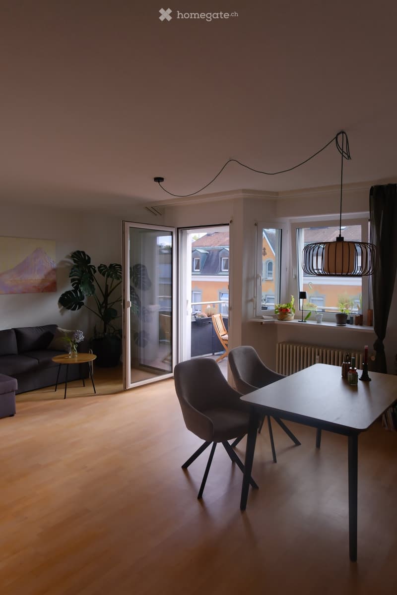 Modern living room with wooden flooring, large windows, and a balcony access. Features a sofa, small round table, two modern chairs, pendant light, and a large plant near the window.