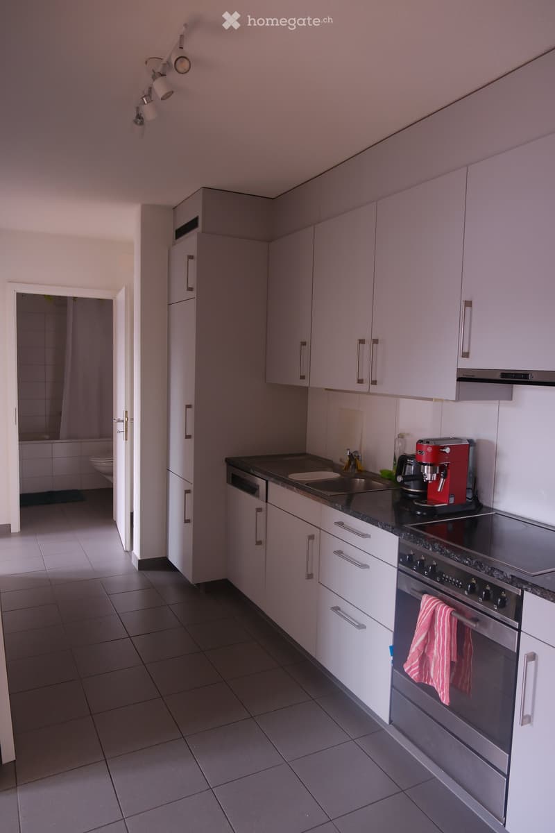 Modern kitchen with white cabinets, stainless steel appliances, and a dark countertop. A red coffee maker and a pink oven mitt are visible on the counter.